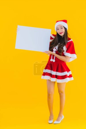 Téléchargez les photos : Portrait belle jeune femme asiatique vêtements de Noël et chapeau sourire avec panneau blanc vide sur fond isolé jaune - en image libre de droit
