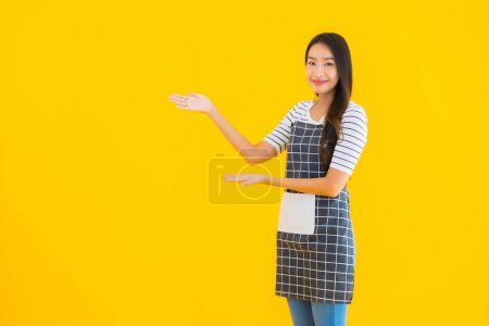 Téléchargez les photos : Portrait belle jeune femme asiatique porter tablier avec sourire heureux sur fond jaune isolé - en image libre de droit