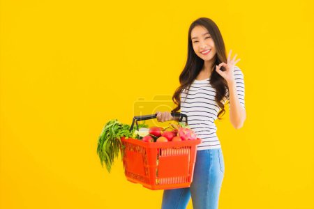 Téléchargez les photos : Portrait belle jeune femme asiatique avec panier d'épicerie panier du supermarché dans le centre commercial sur fond jaune isolé - en image libre de droit