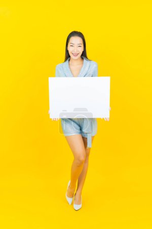 Téléchargez les photos : Portrait belle jeune femme asiatique sourire avec panneau d'affichage blanc vide sur fond jaune - en image libre de droit
