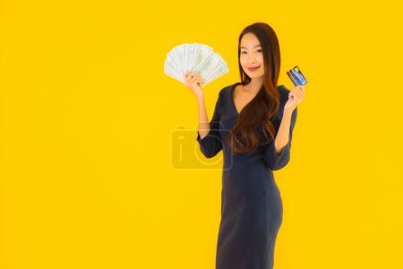 Téléchargez les photos : Portrait belle jeune femme asiatique avec argent comptant et carte de crédit sur fond jaune isolé - en image libre de droit