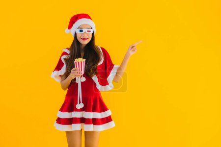 Téléchargez les photos : Portrait belle jeune femme asiatique porter des vêtements de Noël chapeau avec du maïs soufflé et des lunettes 3D prêt pour regarder le film sur fond jaune isolé - en image libre de droit