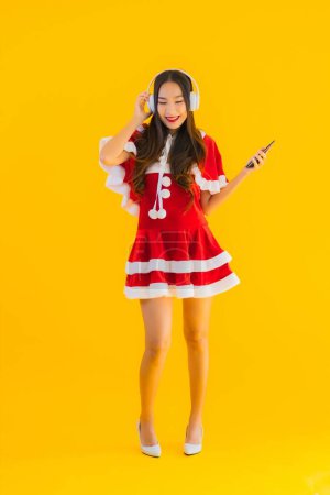 Photo for Portrait beautiful young asian christmas clothes and hat listen for music with headphone and smartphone on yellow isolated background - Royalty Free Image