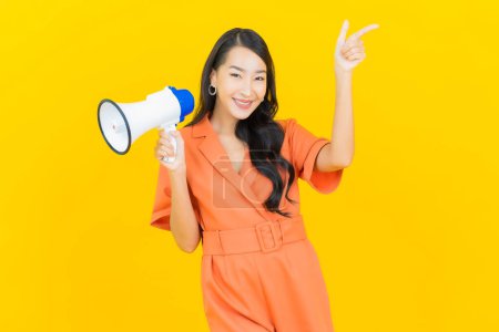 Téléchargez les photos : Portrait belle jeune femme asiatique sourire avec mégaphone sur fond de couleur jaune - en image libre de droit