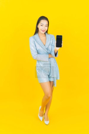 Téléchargez les photos : Portrait belle jeune femme asiatique sourire avec téléphone portable intelligent sur fond de couleur jaune - en image libre de droit
