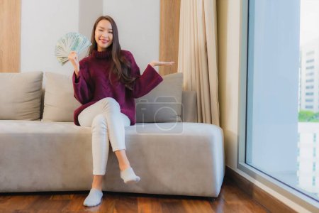 Foto de Retrato hermosa joven mujer asiática con un montón de dinero en efectivo y dinero en el sofá en el interior de la sala de estar - Imagen libre de derechos