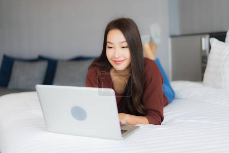 Téléchargez les photos : Portrait belle jeune femme asiatique utiliser ordinateur portable sur le lit dans la chambre à coucher intérieur - en image libre de droit
