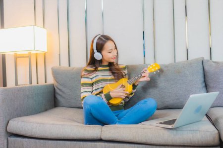 Téléchargez les photos : Portrait belle jeune femme asiatique jouer ukulele sur canapé dans le salon intérieur - en image libre de droit