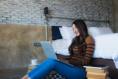 Photo for Young asian woman using laptop computer with coffee cup and book in bedroom interior - Royalty Free Image