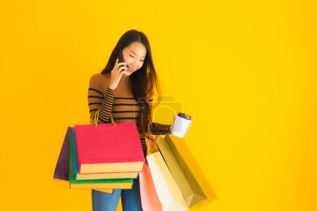 Photo for Beautiful young asian woman use smart mobile phone or cellphone with coffee cup and color shopping bag on yellow isolated background - Royalty Free Image