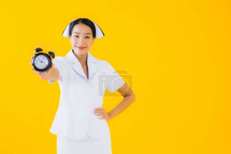 Foto de Retrato hermosa joven asiática mujer tailandesa enfermera mostrar reloj o alarma en amarillo aislado fondo - Imagen libre de derechos