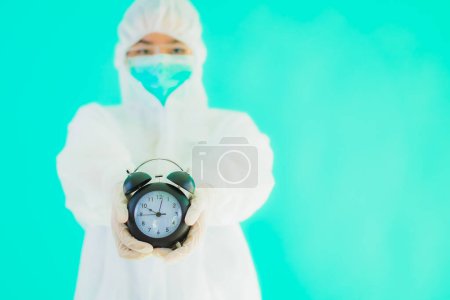 Photo for Portrait beautiful young asian doctor woman wear ppe suite and mask show clock or alarm on blue isolated background - Royalty Free Image
