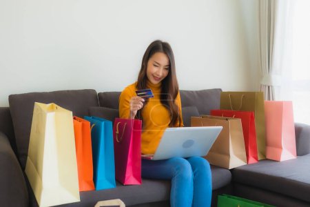 Foto de Retrato joven mujer asiática usando computadora portátil con teléfono móvil inteligente y tarjeta de crédito para compras en línea en la sala de estar - Imagen libre de derechos