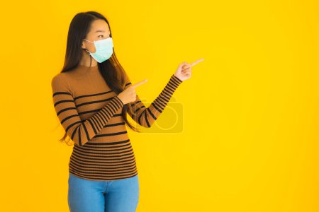 Téléchargez les photos : Portrait belle jeune femme asiatique avec masque dans beaucoup d'action pour protéger de coronavirus ou covid19 sur fond isolé jaune - en image libre de droit