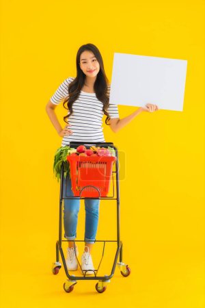 Téléchargez les photos : Portrait belle jeune femme asiatique avec panier d'épicerie chariot et montrer tableau blanc vide sur fond jaune isolé - en image libre de droit