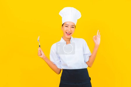 Foto de Retrato hermosa joven asiática mujer usar chef o cocinar uniforme con cuchara y tenedor en amarillo aislado fondo - Imagen libre de derechos