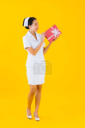 Téléchargez les photos : Portrait belle jeune femme asiatique thai infirmière montrer boîte cadeau rouge sur fond jaune isolé - en image libre de droit