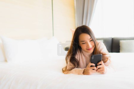 Foto de Retrato hermosa joven mujer asiática con taza de café y teléfono móvil en la cama en el interior del dormitorio - Imagen libre de derechos