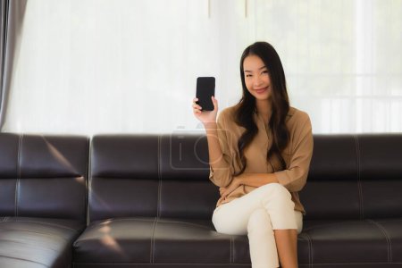 Foto de Retrato hermosa mujer asiática joven utilizar teléfono inteligente móvil o teléfono celular con taza de café en el interior del salón sofá - Imagen libre de derechos