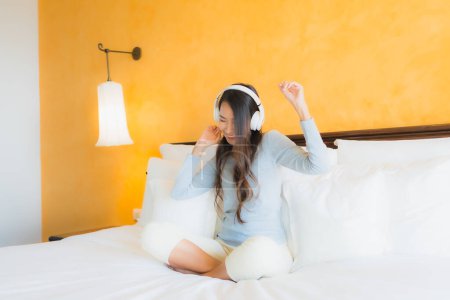 Téléchargez les photos : Portrait belle jeune femme asiatique utilisant un téléphone portable avec casque pour écouter de la musique à l'intérieur de la chambre - en image libre de droit