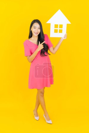 Portrait beautiful young asian woman smile with home sign paper board on yellow color background