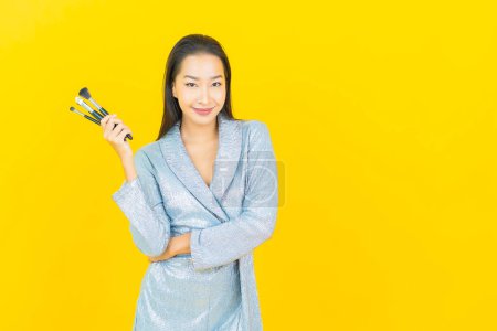 Téléchargez les photos : Portrait belle jeune femme asiatique sourire avec maquillage cosmétique pinceau sur fond jaune - en image libre de droit