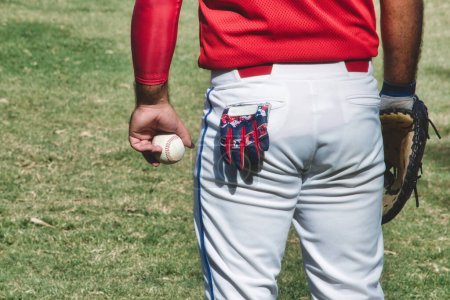 Ein Baseballspieler bei einem Spiel trägt einen Lederhandschuh und hält einen Ball in der Hand, aus seiner Gesäßtasche ragt ein kleiner Handschuh in weißer Hose, der auf dem Rasen steht