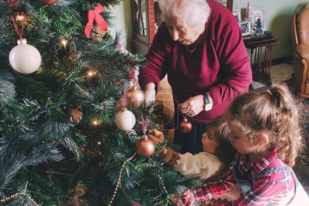 Foto de Un niño y una niña y su abuela decorando el árbol para las vacaciones de Navidad - generaciones más jóvenes y mayores de la misma familia - Imagen libre de derechos