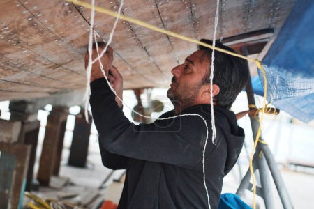 Téléchargez les photos : Un jeune homme d'âge moyen qui travaille sur la coque d'un bateau - mettre du coton dans les fissures entre les planches de bois - en image libre de droit