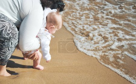 Photo for Barefoot mother and baby daughter on the sea shore on a sandy beach walking close to the surf foam on the waves - Royalty Free Image