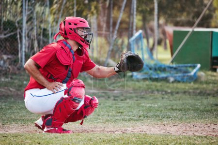 A baseball catcher holding up a leather glove crouching  down on the grass pitch