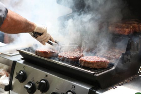 Photo for Close-up of a man's hand holding a pair of tongs, turning beef steaks on a smoky barbeque - Royalty Free Image