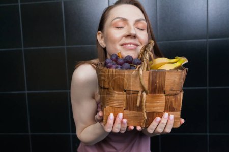 Foto de Una chica con una cesta tejida de frutas saludables, azulejos oscuros en el fondo. Concepto de comida saludable y estilo de vida. desintoxicación y dieta limpia. - Imagen libre de derechos