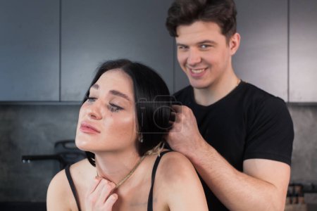 Téléchargez les photos : Domestic violence concept. A man strangles a woman with rope at home. A frightened young woman cries, a man stands and smiling behind her. family problems. - en image libre de droit