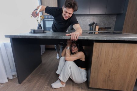 Téléchargez les photos : Woman sitting and crying under kitchen table scared of man attacking with knife. family problems. Domestic violence concept. - en image libre de droit