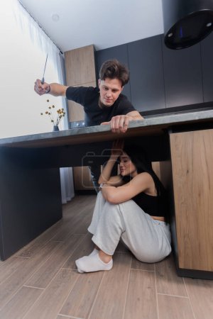Téléchargez les photos : Woman sitting and crying under kitchen table scared of man attacking with knife. family problems. Domestic violence concept. - en image libre de droit