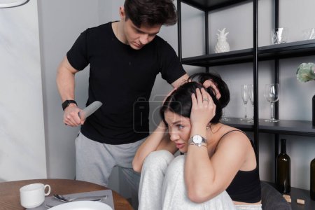 Téléchargez les photos : Woman sitting and crying at kitchen table scared of man attacking with knife. family problems. Domestic violence concept. - en image libre de droit
