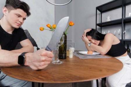 Téléchargez les photos : Woman sitting and crying at kitchen table scared of man attacking with knife. family problems. Domestic violence concept. - en image libre de droit