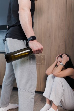 Téléchargez les photos : Frightened woman sitting at the corner and crying scared of drunken husband or boyfriend with a bottle in his hand. Drunkenness in the family. family problems. The concept of domestic violence. - en image libre de droit