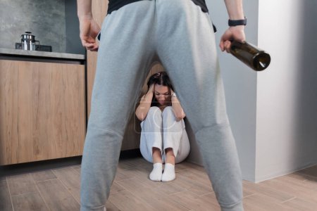 Téléchargez les photos : Frightened woman sitting at the corner and crying scared of drunken husband or boyfriend with a bottle in his hand. Drunkenness in the family. family problems. The concept of domestic violence. - en image libre de droit