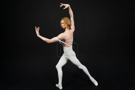 Photo for Young athletic professional ballet dancer with a bare torso and white dance tights is in perfect shape and posing with bright yellow slippers in his hands over a black background. - Royalty Free Image