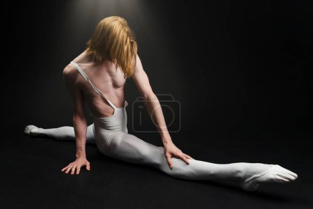 Photo for Young athletic professional ballet dancer with a bare torso and white dance tights is in perfect shape, performing and doing splits over a black background. - Royalty Free Image