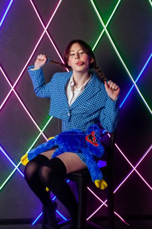 Photo for Portrait of a cute girl with drawn tears, dressed in a blue plaid jacket and skirt with knee high socks, sitting in a neon room pulling pigtails and showing tongue. - Royalty Free Image