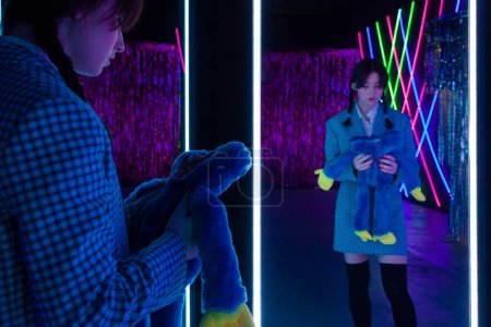 Photo for A cute girl with drawn tears, dressed in a blue plaid jacket and skirt with knee high socks, holds soft toy and looks mysteriously at herself in the mirror in a neon-lit room. - Royalty Free Image