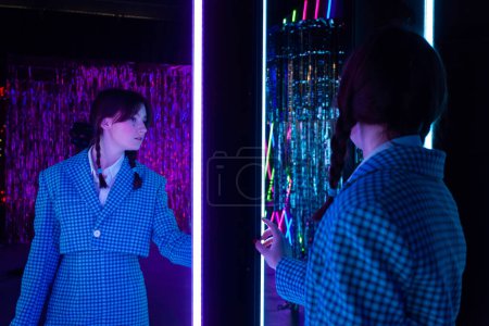Photo for A cute girl with drawn tears, dressed in a blue plaid jacket and skirt with knee high socks, looks mysteriously at herself in the mirror in a neon-lit room. - Royalty Free Image