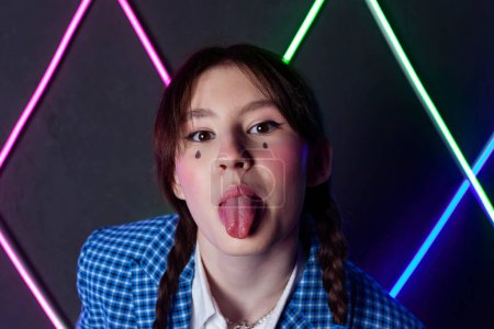 Photo for Portrait of a cute girl with drawn tears, dressed in a blue plaid jacket, shows a tongue in a neon lit room. - Royalty Free Image