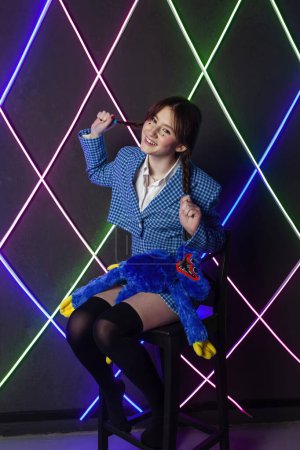 Photo for Portrait of a cute girl with drawn tears, dressed in a blue plaid jacket and skirt with knee high socks, sitting in a neon room, smiling and pulling pigtails. - Royalty Free Image