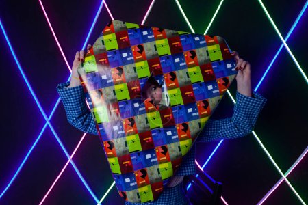 Photo for Portrait of a cute girl with drawn tears, dressed in a blue plaid jacket,  peeking out from a cutout in a sheet of glitter wrapping paper in a neon lit room. - Royalty Free Image