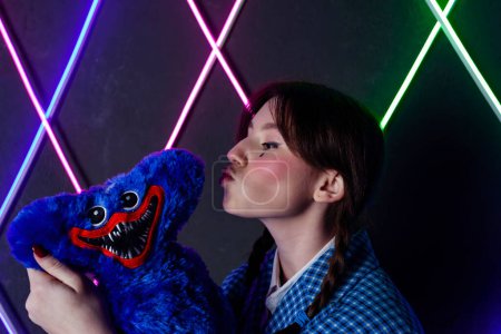 Photo for Portrait of a cute girl with drawn tears, dressed in a blue plaid jacket, kissing a soft toy in a neon lit room. - Royalty Free Image