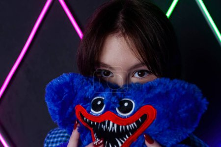 Photo for Portrait of a cute girl with drawn tears, dressed in a blue plaid jacket, holding and peeking out from behind a soft toy in a neon lit room. - Royalty Free Image